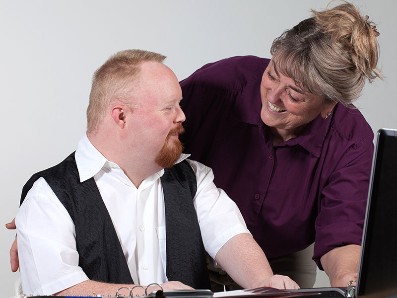 A woman who is standing up leans over to assist a seated man with using a computer. Both people are smiling.