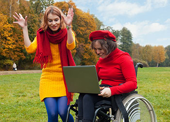 Two women wearing colorful clothing are outside in a field on a sunny autumn day. The woman on the left is gesturing with her hands, and the woman at the right is sitting in a wheelchair with a portable computer in her lap. She is looking at the screen.